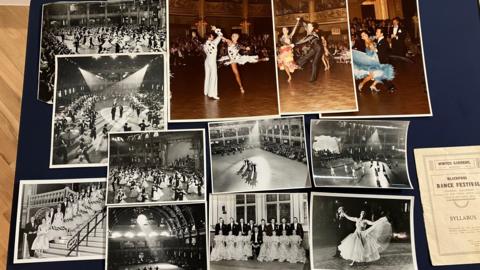 A collection of photographs on a blue board. The majority are in black and white, showing people dancing in large ballrooms. There are three in faded colour towards the top right. Towards the bottom right, an old syllabus for Blackpool Dance Festival can be seen