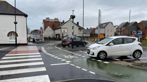 A white small car leaving the roundabout and grey small car entering the roundabout. They are crossing over the green painted cycle lane and zebra crossing which are empty in the picture. The area is built up with housing and there is a pub in the background