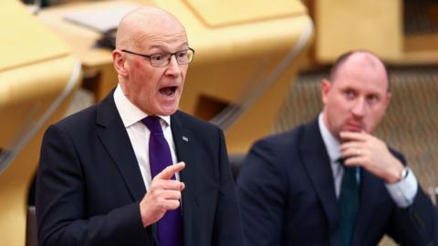 First Minister John Swinney on his feet in the chamber gesticulating with Health Secretary Neil Gray sitting pensively in the background.