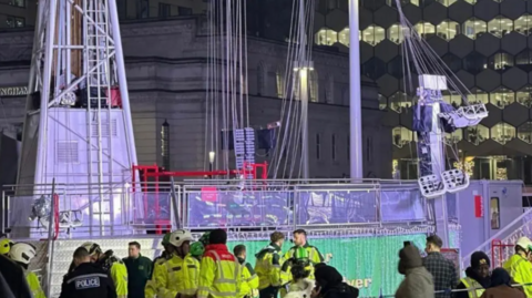 Emergency services and other people gather next to a fairground ride. A section of the ride, where people sit, appears to have crashed into a post on the right.