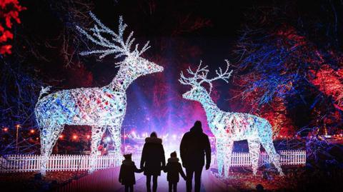 A family of four with warm coats and hats stand in front of a pair of giant white illuminated reindeers in the grounds of Dunham Massey with orange and blue lights and spotlights on lighting up trees behind them.    