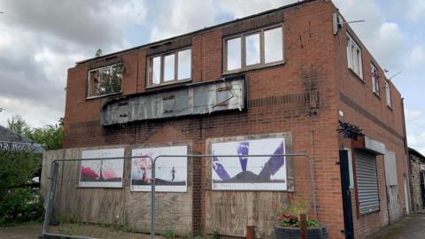 A derelict red brick building with broken windows is boarded up with a wire fence around it