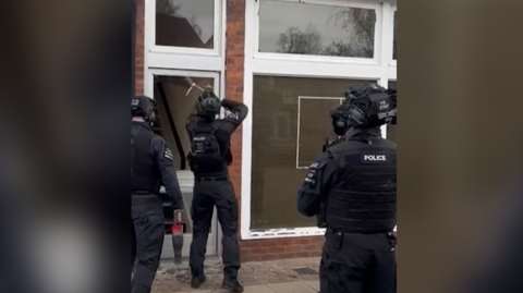 Three police officers in black tactical equipment are stood outside a brick building with large white window frames. The officer closest to the building uses some equipment to break glass around the door.