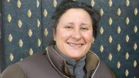 A woman with short black hair smiles at the camera. Behind her is a curtain with a gold leaf-like design on a dark background. She wears a puffy brown jacket over a grey fleece top.