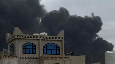 A black cloud of smoke hovers above a building with blue windows