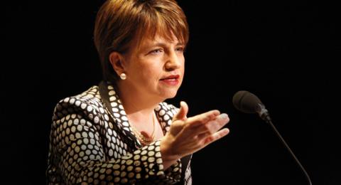 A woman with short dark hair and a dotted top speaks into a microphone 