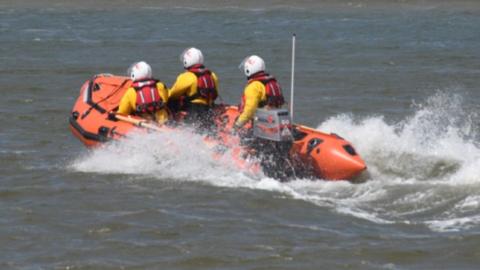 An orange lifeboat belonging to the RNLI showing three RNLI crew members on the sea, on the way to an incident. 
