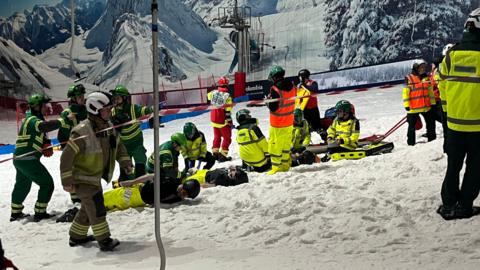 Emergency responders in high-visibility jackets and helmets gather on an indoor snow slope, treating two false models lying on the ground while others prepare a stretcher nearby. A ski lift and a mural of snowy mountains are visible in the background, and red safety barriers mark the area.
