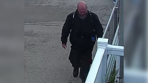 A man wearing a dark green bomber jacket, black cargo trousers and a black backpack. He is bald with a beard and is walking with his head facing the ground. There is a white fence to the right of the image. 