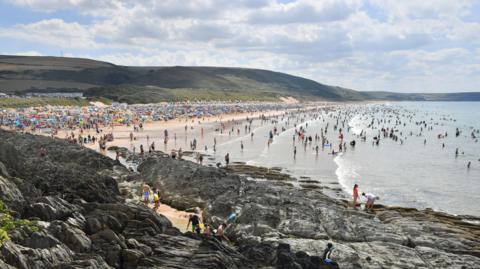 Woolacombe beach on a sunny day is packed with people and the sea is full of people wading and swimming. The grassy headland stretches down the coast and there are cars lined along a road on it. In the foreground there are rocks which a few people are standing on.