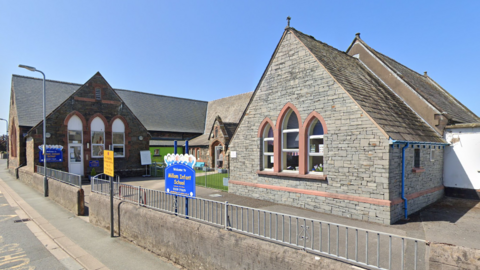 A Google Street View screenshot of Millom Infant School. It is off a main road and has Cumbrian slate cladding at the front of the building. It has a small court yard at the front of the entrance.