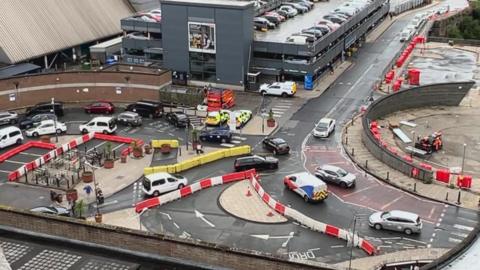 A picture of the drop-off system at Leeds Railway Station. There are vehicles queuing to access the loop, with several taxis parked up. There is a car park and buildings in the background.