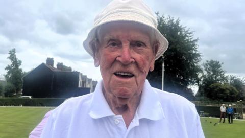 An elderly man wearing a white bucket hat and a white shirt smiles for the camera with a lawn bowls green and houses pictured behind him and two people stand over some bowls.