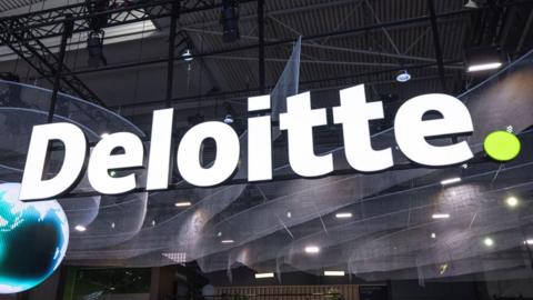 A large Deloitte sign in white letters with a small green full stop at the end. The large lighted sign is hanging over a large hall floor and there is a pixelated globe on the left of the picture.