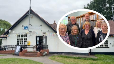A white building with multicoloured bunting strung up in front. The building has a sign that reads 'Anlaby Park Library' and 'Run by the community for the community'. The library has a black front door which is open, and has red steps and a disabled ramp leading up to it. Above, the sky is cloudy and grey and you can see green trees in the background. Below is a path and green grass with white flowers on. To the right is a white oval featuring a group picture of five women and two men in a library setting, with a long brown bookshelf behind them filled with books.