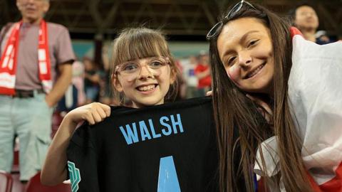 A young girl smiles as she holds up the reverse side of a black England away shirt with the name Walsh and a number 4 printed on the reverse. An adult - presumably her mum - has an England flag wrapped around her shoulder as she leans into the shot.