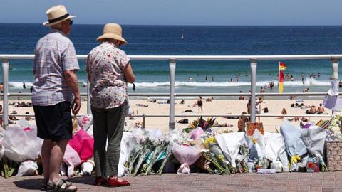 Two people stand in front of floral tributes on a paved path with a beach in the background