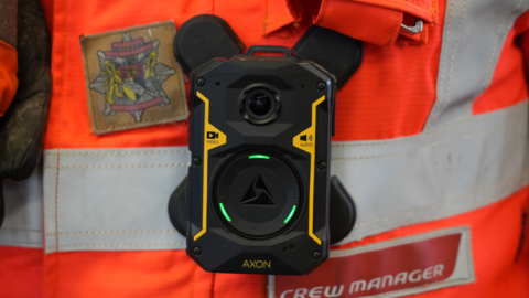 A close-up of a body-worn camera, positioned on an orange and reflective silver top. 
