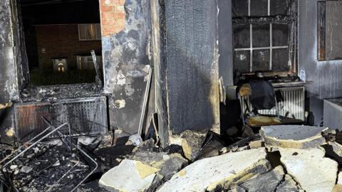 A burnt out room with bits of blackened debris. The wall can be seen split into pieces and scattered across the ground. The windows are also blown out. 