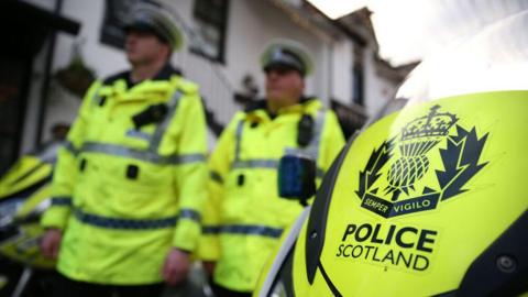 Two police officers in fluorescent jackets are blurred. The image focuses on the front of a police motorbike with Police Scotland written on it and a crest which reads "Semper Vigilo".