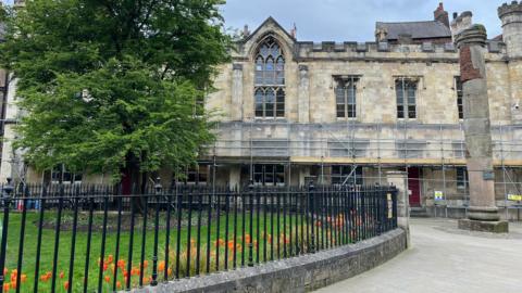 An old building with large ornate windows and a crenellated top, with scaffolding around the bottom of it. An area of grass with railings around it in front of the building with orange tulips in a bed edging the grass.