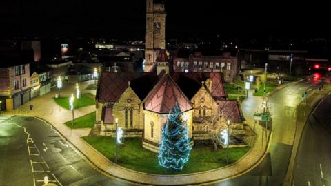 An aerial view of the tree decorated with blue lights at night and there are no people in the street. It is behind a church with clock and near a road. The snap is from 2023.