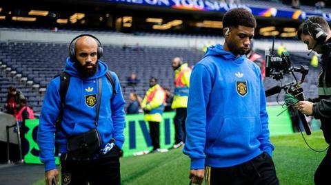 Amad Diallo and Bryan Mbeumo, wearing blue Manchester United hoodies, walking on the side of the pitch on their way to the away dressing room at an empty Tottenham Hotspur Stadium