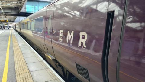 An intercity purple coloured train has the letters EMR on the side. It is pulled in to a platform on Sheffield railway station