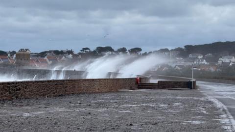 Large waves crash over a seawall in Guernsey during stormy weather brought by Storm Benjamin on Thursday. Parts of the road next to the wall have puddles and surface water. Thick grey clouds are in the sky.