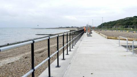 Concrete path with metal railing on left, with pebble beach and sea