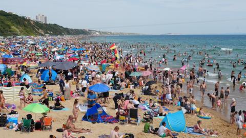 A beach is filled with thousands of sunbathers, parasols and tents as crowds flock to the beach in the sunshine. Many are paddling and swimming in the sea. red and Yellow lifeguards flags can be seen standing in the sand.