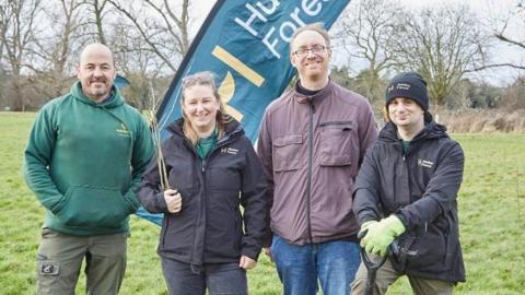 Volunteers and members of the community standing in a field with trees in the background, wearing coats and warm clothing. One volunteer is holding a shovel and has one foot rested on it. 