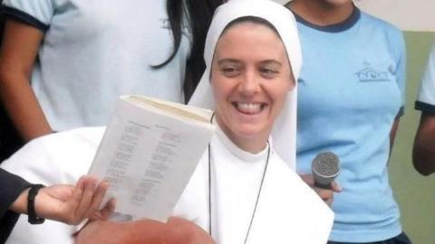 Shows a nun in a white habit and veil smiling. Someone is holding a booklet up for her to see.