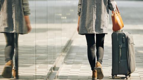 A woman with her back to the camera wheeling a suitcase down the street.