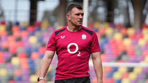 Ben Spencer is wearing a red England training shirt and standing on the pitch during a pre-match warm-up ahead of the game with Argentina in July.