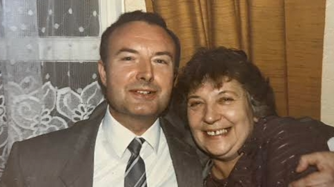 An old photograph of Mr Hill with a woman. Both are smiling at the camera. Mr Hill has short dark hair and is wearing a grey suit and a grey tie, while the woman is in a dark blouse.