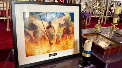 A picture is displayed showing an image of soldiers with shields emerging from fire. In the bottom right of the picture is a white candle with ribbon wrapped around it in the blue and yellow colours of the Ukrainian flag.