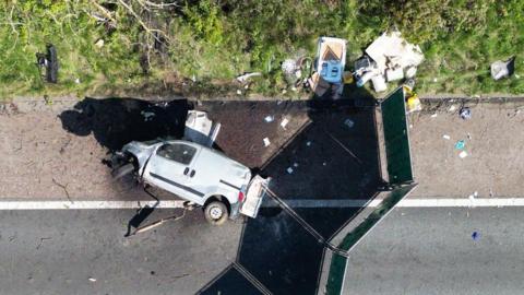 An aerial drone shot of the van, lying on its side on the hard shoulder of the motorway. Its bonnet is badly damaged and the passenger side front wheel has been torn off. There is other debris littering the grass verge, and a screen has been put up around the scene. 