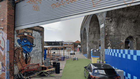 A metal shutter is half raised. Inside the venue there is a car parked on some artificial grass. To the left is a canopy with benches and tables beneath it. A series of large bricked archways are positioned on the right of the venue with a centre stage at the back.