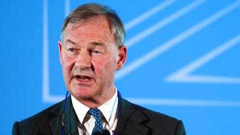 Rupert Lowe is wearing a dark business suit, a white shirt, patterned blue tie and has a pair of glasses hanging round his neck. He is standing at a podium with a microphone. The background is a bright blue presentation screen with abstract white geometric lines.