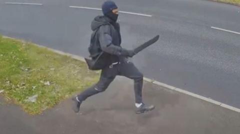 man wearing all back runs across grass verge holding a machete