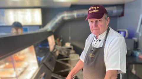 A man stands behind a row of chrome-coloured deep friers and a glass-fronted counter at a fish and chip shop. He is wearing a grey apron and burgundy cap, both with "Railway St Fisheries" logos on them, and a white shirt.