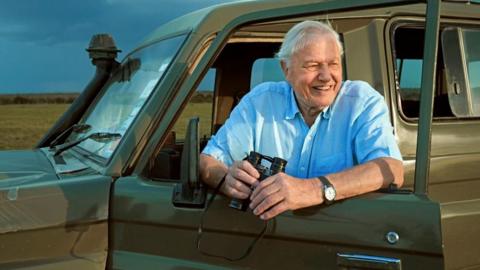 David Attenborough leaning out of jeep with binoculars in his hand