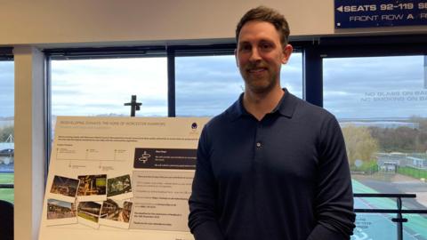 A man with short brown hair and a dark blue long-sleeved polo shirt stands in front of a sign promoting the redevelopment of Sixways Stadium.