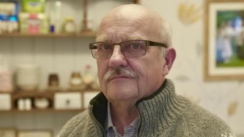 Gary Lovatt looking into the camera with a straight face. He is mostly bald with white hair on the side of his head and a white moustache. He is wearing rectangular glasses and is wearing a cable-knit grey cardigan over a polo shirt. He looks to be sitting in a kitchen which is slightly blurred. There is a large shelf behind him with bowls and Tupperware on it. 