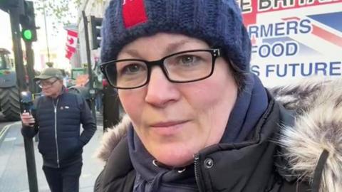 Anna Longthorp stands on the pavement in London with tractors on the road and a banner behind her.  She wears glasses and a blue woollen hat and hooded top and a black coat with fur trim.