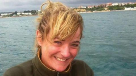 A woman with blonde air and a brown sweater with a yellow rim smiles at the camera, with the sea in the background