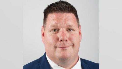 Nick Housden headshot. He is wearing a navy blazer and white shirt, and has short brown hair which is gelled up. He is slightly smiling.