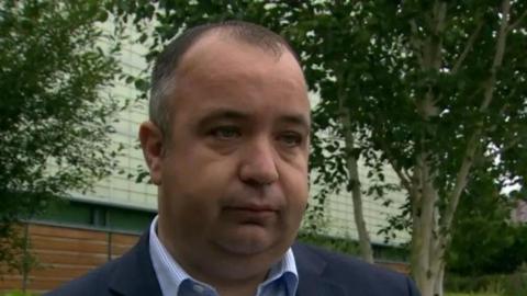 Brian Tierney has a navy suit and blue shirt on. He has thin, receding brown and grey hair. Behind him is a white building and two green tree's. 