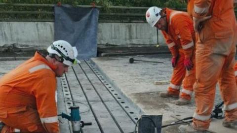 Three men, in orange high-vis jumpsuits, working on a viaduct. You can see concrete floor. One man is kneeling down wearing a white hard-hat and just two other peoples legs can be seen. There is machinery on the ground.